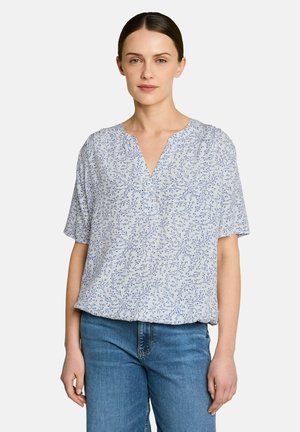 Kobieta w jasnoniebieskiej bluzce z krótkim rękawem z małym kwiatowym wzorem i niebieskich dżinsach, stojąca na jednolitym tle.