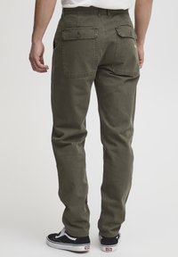 Pantalon cargo vert olive en coton, avec deux poches arrière boutonnées et une coupe droite. Porté avec des baskets noires.