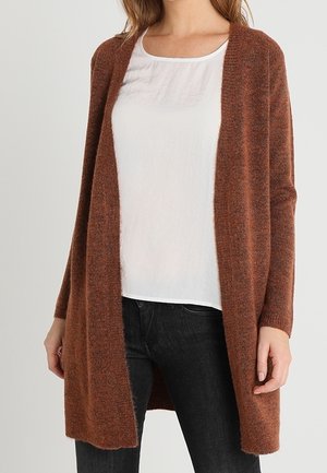 Femme portant un cardigan marron ouvert devant sur un chemisier blanc, associé à un jean noir, debout devant un fond uni.