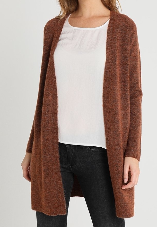 Femme portant un cardigan marron ouvert devant sur un chemisier blanc, associé à un jean noir, debout devant un fond uni.