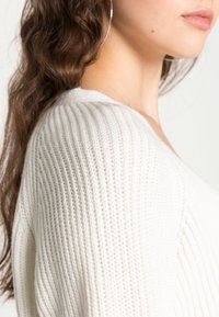Suéter ligero, de canalé, en crema con cuello en V. Detalles en primer plano de su textura suave y patrón de punto abierto visibles.