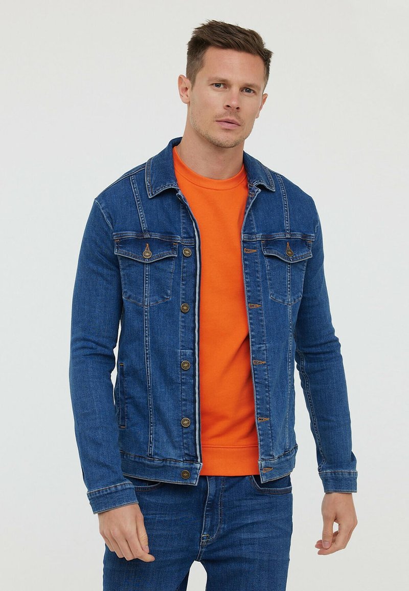 Veste en denim bleu avec deux poches poitrine, fermeture à boutons et coutures contrastantes, superposée sur un sweat-shirt orange. Texture lisse.