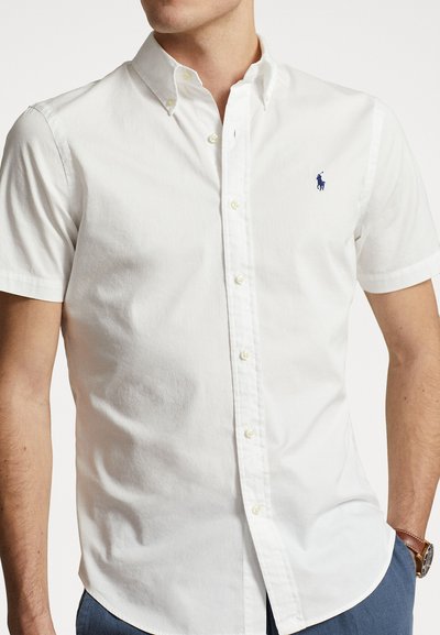 Chemise blanche en coton à manches courtes avec un logo brodé bleu sur la poitrine gauche, dotée d'un col classique et d'un ourlet droit.