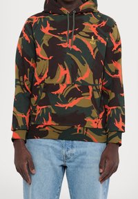 Sweat à capuche avec un motif camouflage multicolore incluant des nuances de vert, marron et rouge, doté d'une poche kangourou à l'avant et de poignets côtelés.