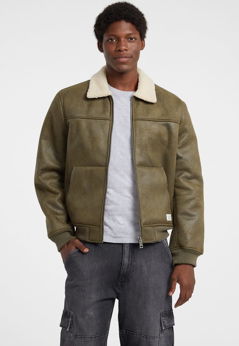 Giacca bomber verde oliva con colletto in shearling, chiusura con zip e polsini a coste. Presenta due tasche frontali e una finitura liscia e testurizzata.