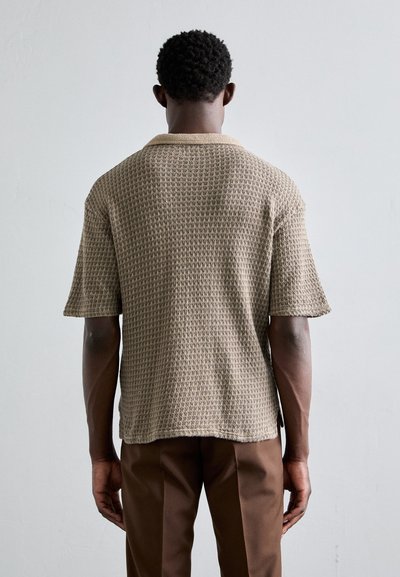 Camisa de punto de manga corta en beige con un patrón de waffle texturizado y un cuello suave, combinada con pantalones marrones.