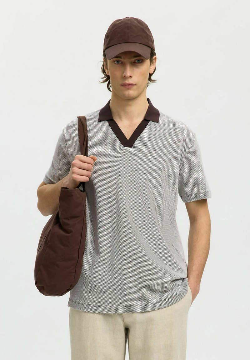 Jeune homme portant une casquette marron, une chemise grise à manches courtes avec un col foncé, un pantalon beige et un sac à bandoulière marron.