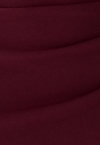 Bordový textil s hladkou štruktúrou, jemným sklápaním a miernymi vlnami, ktoré vytvárajú hĺbku prostredníctvom variácií svetla a tieňa.