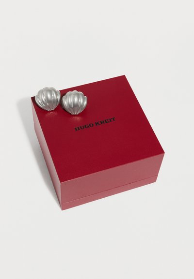 Δύο ασημένια σκουλαρίκια σε σχήμα κοχυλιού, τοποθετημένα πάνω σε ένα κόκκινο τετράγωνο κουτί με την επιγραφή "HUGO KREIT" στο καπάκι.