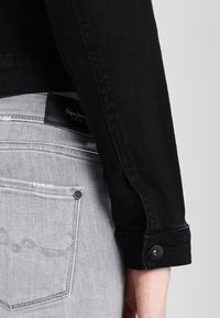 Svart denimjacka med figursydd design, utrustad med knappmanchetter och en strukturerad yta, kombinerad med ljusgrå denimjeans.