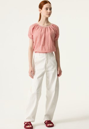 Haut à manches bouffantes à carreaux rouges et blancs avec encolure élastique, associé à un pantalon blanc ample et des sandales tressées rouges. Fond uni.