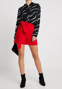 Žena v černo-bílé blůze s abstraktním vzorem, s červenými šortkami s vysokým pasem a předním zavazováním, černými kotníkovými botami a držící černý psaníčko.