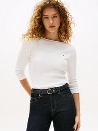 Weißes Langarmoberteil mit Scoop-Ausschnitt, kombiniert mit dunkelblauen Jeans und einem schwarzen Gürtel mit einer silbernen Schnalle. Minimale Markierung sichtbar.