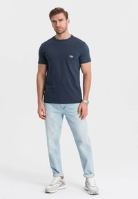 Námorně modré bavlněné tričko s náprsní kapsou a logem. Vydáváno s světle modrými džínami s odřeným vzhledem a šedými sportovními botami.