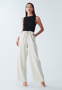 Melns teksturēts krekls bez piedurknēm kopā ar gaiši bēšām platām bikšu kājām, kurām ir aukliņu josta. Modele stāv uz vienkārša fona.