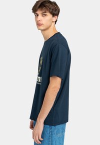 Camiseta de algodón azul marino con mangas cortas y cuello redondo. Presenta un logo y texto dorado en la parte frontal. Corte casual y relajado.