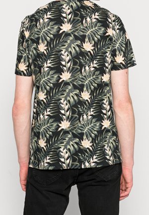 Homme portant une chemise noire à manches courtes avec des motifs de feuilles et de fleurs tropicales vertes et beiges, associée à un pantalon noir, vu de dos.