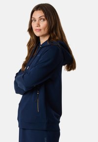 Navy blauwe hoodie van zacht materiaal, met een voorzak met een gouden ritsdetail, een verstelbare trekkoord capuchon en ribgebreide manchetten.