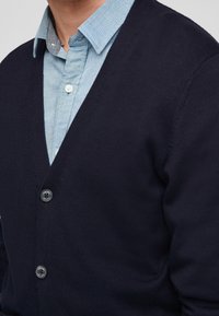 Námořnický pletený cardigan s výstřihem do V, třemi knoflíkovými zapínáními a ribbovanou texturou, nošený přes světle modrou košilovou košili.