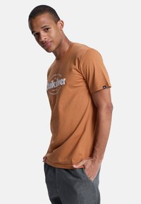 Braunes T-Shirt mit kurzen Ärmeln aus Baumwolle, mit einem weißen Logo auf der Vorderseite und einem kleinen schwarzen Etikett am Ärmel. Glatte Textur.