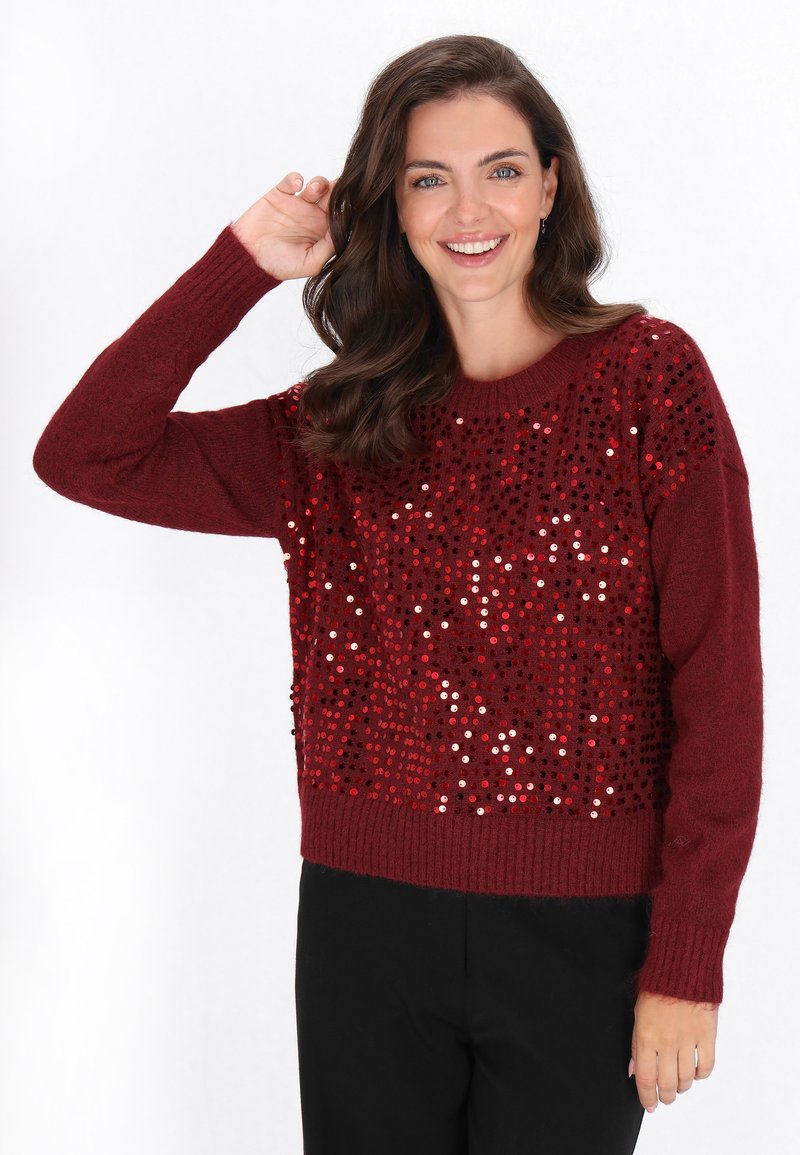 Donna sorridente con lunghi capelli castani, che indossa un maglione bordeaux con paillettes rosse, alzando una mano vicino alla testa, in piedi contro uno sfondo bianco.