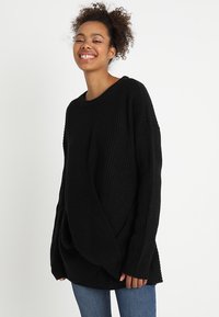 Schwarzer Strickpullover mit lockerer Passform, gerippter Textur, weitem Ausschnitt und fallenden Schultern, kombiniert mit blauen Jeans.