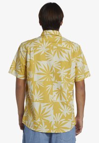 Camicia a maniche corte gialla con fantasie di foglie bianche, caratterizzata da un design con colletto e orlo curvo, realizzata in tessuto leggero.