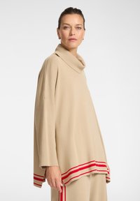 Maglione beige con collo a scialle e maniche ampie, caratterizzato da una striscia orizzontale rossa lungo l'orlo. Tessuto morbido e testurizzato, vestibilità ampia.