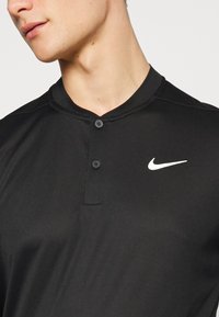 Černé polo tričko značky Nike s límečkem, třemi knoflíky a bílým logem na levé straně hrudi. Hladká textura a přiléhavý střih.