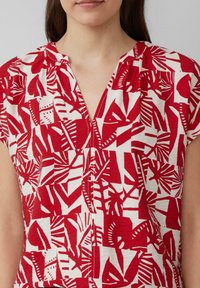 Haut rouge et blanc à manches courtes avec un col en V, présentant un motif géométrique de feuilles et un tissu texturé. Coupe ajustée avec des détails de plis.