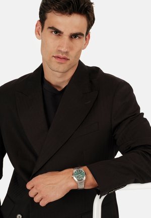 Sort dobbeltradet blazer, struktureret stof, båret over en mørk top. Sølvfarvet armbåndsur med grøn skive, med en linket metalrem.