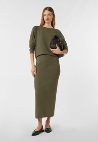 Dzianinowy sweter w oliwkowej zieleni z okrągłym dekoltem, zestawiony z dopasowaną spódnicą do kolan. Modelka trzyma ciemnobrązową torebkę, ma na sobie czarne baleriny.