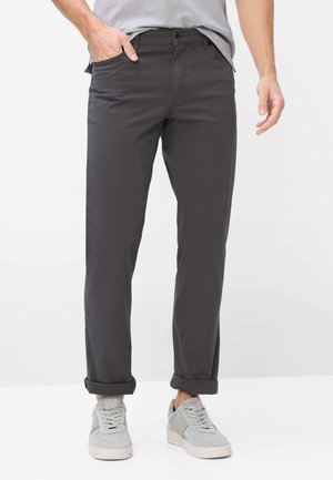 Mand iført mørkegrå bukser med opkrogede ben, grå sneakers og en lysegrå t-shirt, står med den ene hånd i lommen.