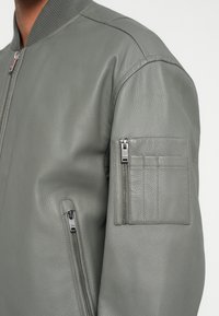 Harmaa nahkainen bomber-takki, jossa on resorikaulus, vetoketjulliset sivutaskut ja vetoketjullinen hihatasku, jossa useita korttipaikkoja. Rakenteellinen viimeistely.