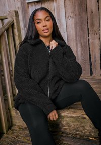 Mustat, pörröiset vetoketjulliset takit, joissa on kaulus ja teksturoitu fleece-materiaali. Malli istuu puuporrastuksilla, pukeutuneena mustiin leggins-leggingsiin.