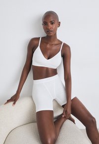 Hvit bralette og boyshort-sett i glatt stoff, med tynne stropper og en tettsittende design. Modell poserer på en teksturert lys grå overflate.