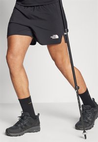Sorte idrettsshorts med en logo, kombinert med sorte tursko og mørke sokker, mens man holder en turstang.