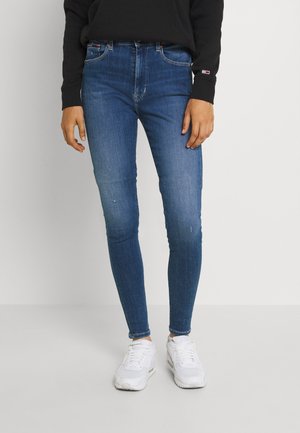 Persoon draagt strak zittende blauwe jeans, witte sneakers en een zwart sweatshirt, staand tegen een effen lichte achtergrond.