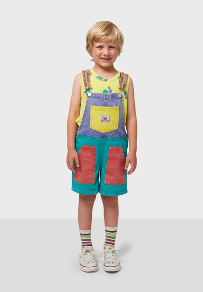 Multicolored dungarees with yellow, purple, and teal sections; features large red pockets and adjustable straps. Worn with a yellow shirt and striped socks.