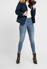 Kvinna som bär en marinblå pufferjacka med fuskpäls på huvan, vit tröja, urblekta blå skinny jeans och bruna ormskinns ankle boots, poserar.