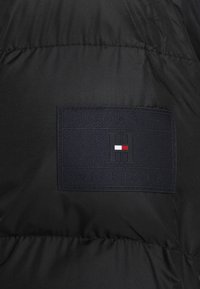 Giubbotto imbottito nero con un patch logo testurizzato nero che riporta la scritta "TOMMY HILFIGER" e una "H" stilizzata accentuata in rosso e bianco.