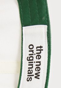 Grønt stoffrem med en hvit etikett som viser "the new originals" i svart tekst. Teksturoverflate med hvit underside.