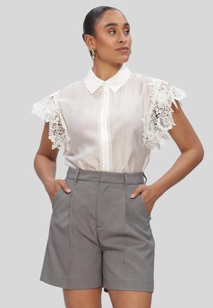 Frau mit Händen in den Taschen, trägt eine weiße Bluse mit Spitzen-Blumenärmeln und graue hoch taillierte Shorts, schaut zur Seite.