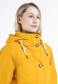 Chaqueta amarilla impermeable con capucha, con botones a presión, cordones ajustables y una textura suave. Diseñada con acentos prácticos.