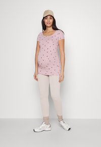 Light pink maternity top with short sleeves, small black bird patterns, paired with light beige leggings and silver athletic shoes.