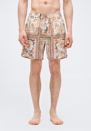 Muž oblečený v šortkách s farebnými, zložitými vzormi zobrazujúcimi slnka, zvitky a ornamentálne motívy, stojaci naboso na bielom pozadí.