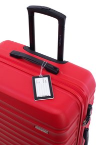 Valise en polycarbonate rouge avec un design strié, une poignée noire extensible et une étiquette de bagage attachée. Elle dispose d'une fermeture à glissière et d'un logo en métal.
