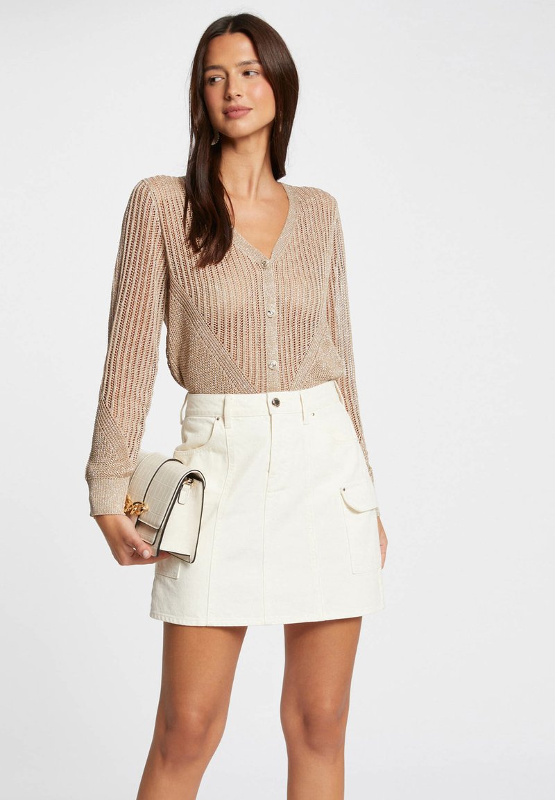 Beige, gestrickte Strickjacke mit V-Ausschnitt und Knopfverschluss, kombiniert mit einem weißen Minirock mit Fronttaschen. Hält eine strukturierte Clutch.