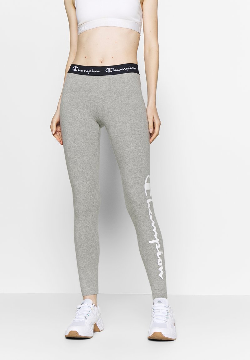 Szare legginsy sportowe z czarnym pasem na biodrach z napisem "Champion". Materiał jest gładki i dopasowany, z dużym białym logo na lewej nodze.