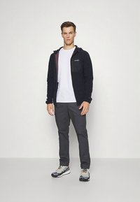 Tumši zila flīsa jaka ar kontrastējošu iekšējo odere, kombinēta ar baltu T-kreklu un pelēkām biksēm. Modelis valkā pelēkus sporta apavus.